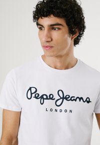 Bijela pamučna majica s crnim kursive natpisom "Pepe Jeans" i "LONDON" ispod. Klasičan okrugli izrez i kratki rukavi.