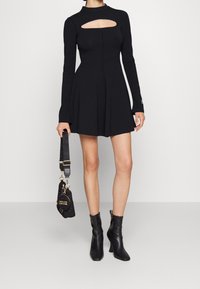 Svart långärmad klänning med figursydd överdel och utstansad detalj. Modellen håller en liten svart handväska med guldfärgade detaljer och har på sig svarta ankelboots.