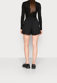 Schwarze Shorts mit elastischem Bund, die eine lockere Passform und einen weichen Stoff aufweisen. Kombiniert mit schwarzen klobigen Schuhen, von hinten betrachtet.