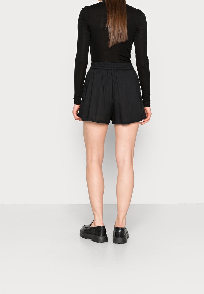 Schwarze Shorts mit elastischem Bund, die eine lockere Passform und einen weichen Stoff aufweisen. Kombiniert mit schwarzen klobigen Schuhen, von hinten betrachtet.