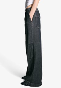 Črne široke denim hlače z elastičnim pasom, stranskimi žepi in vidnimi šivi. Tkanina ima gladko teksturo.