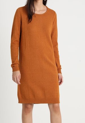 Femme portant une robe en maille couleur rouille, à manches longues et longueur au genou, avec des poignets et un ourlet côtelés, debout devant un fond uni.