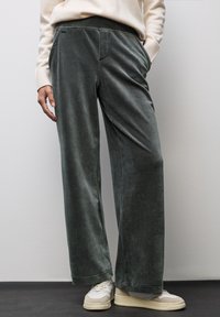 Pantalons larges en velours vert avec une texture lisse, dotés de poches biais et d'une coupe décontractée, associés à des baskets blanches et beiges.