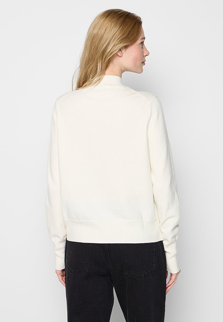 Frau mit blonden Haaren, die einen weißen Langarmpullover und schwarze Jeans trägt, von hinten vor einem schlichten hellen Hintergrund gezeigt.