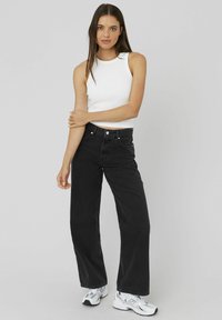 Jeans noirs à jambe large avec une taille haute, dotés de cinq poches et d'un ourlet brut, associés à un débardeur blanc ajusté et des baskets blanches.