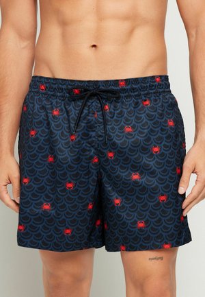 Marineblauwe zwemshorts met schelpmotieven en kleine rode krabbenprint, gedragen door een persoon zonder shirt met ontspannen handen langs het lichaam.