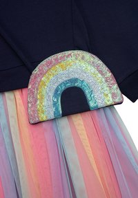 Dunkelblaue Kapuzenjacke mit einem Pailletten-Regenbogen-Patch; mehrlagiger Tüllrock in Pastellfarben wie Rosa, Blau und Lavendel mit vertikalen Streifen.