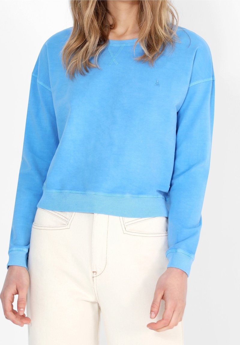 Felpa blu con scollo a girocollo, polsini e orlo a costine, e dettagli di cucitura minimi. Indossata sopra pantaloni chiari a vita alta.