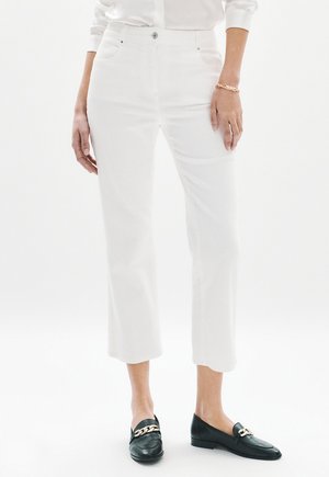 Frau trägt weiße, hoch taillierte, gekürzte Hosen, ein goldenes Armband und schwarze Loafers mit goldenen Kettendetails, steht vor weißem Hintergrund.