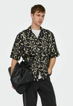 Jonge man die een zwart-beige bloemenprint kort mouw overhemd draagt, zwarte spijkerbroek, houdt een zwarte leren tas vast, kijkt iets opzij.