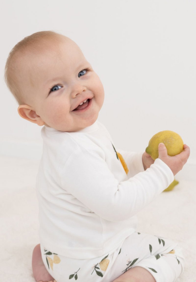 Weißes Langarmshirt, gemusterte Leggings mit Zitronenmotiv. Baby sitzt, hält eine Zitrone und lächelt. Weicher Hintergrund, hellfarbener Boden.