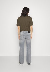 Olijfgroene katoenen t-shirt gecombineerd met lichtgrijze flared jeans met achterzakken en een zwart merklabel. Schoenen zijn zwart-wit.