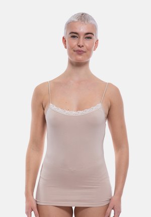 Woman with short blonde hair wearing a beige camisole with thin straps and lace trim, standing against a white background.
