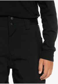 Osoba v čiernych nohaviciach s dvojitým gombíkom a suchým zipsom, v čiernom tričku s dlhými rukávmi, s rukou položenou blízko bedra.