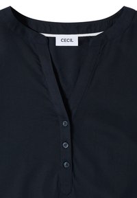 Chemise noire avec encolure ronde et trois boutons, avec une étiquette blanche portant le nom de la marque « CECIL » à l'intérieur du col.