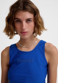 Blue sleeveless top made of smooth fabric with a rounded neckline and debossed "hummel PERFORMANCE" logo on the front. Silver chain necklace visible.