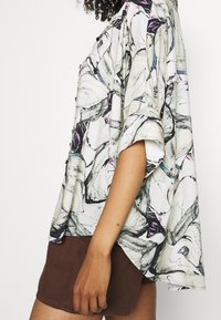 Camisa de manga corta en tejido ligero, con un patrón floral en blanco, gris y toques de púrpura, combinada con pantalones cortos marrones.