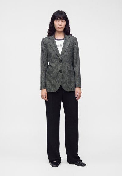 Femme aux longs cheveux foncés portant un blazer gris à carreaux, un t-shirt blanc avec un motif graphique, un pantalon large noir et des chaussures noires pointues, debout devant un fond blanc.