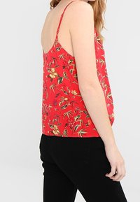 Rød blomster tanktop med tynde stropper, der har et levende blomster mønster i gul og grøn, lavet af letvægtsstof. Sorte bukser synlige.