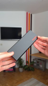 Mustast ristkülikukujulisest power bank'ist, millel on matt viimistlus, millel on kõrgendatud punane "HUGO" tekst, hoituna kahe käe vahel lihtsa siseruumi taustal.