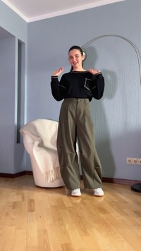 Un haut noir à manches longues avec des accents blancs, un pantalon large taille haute vert olive et des baskets blanches, devant un mur bleu clair.