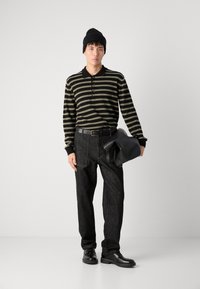 Polo cu mânecă lungă în dungi negre și măslinii, blugi negri, curea neagră, pantofi negri, căciulă, ținând o geantă neagră.