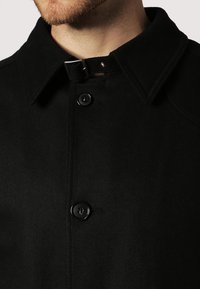Cappotto in misto lana nero con colletto classico, bottoni neri e una cinghia di chiusura. Il tessuto ha una texture liscia con un design strutturato.