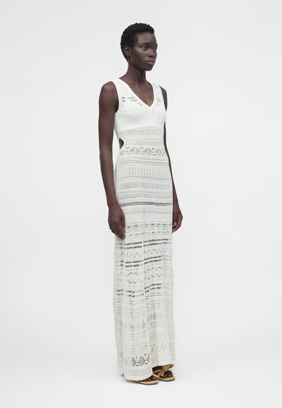 Γυναίκα με λευκό άμανικο μακρύ πλεκτό φόρεμα με ανοίγματα στο πλάι και κίτρινα σανδάλια με κορδόνια, στέκεται μπροστά σε ένα απλό λευκό φόντο.