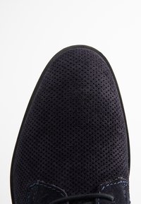 Chaussure en suède bleu foncé avec une texture perforée. Bout arrondi et semelle en caoutchouc noir. De fines coutures bleues soulignent la zone des lacets.