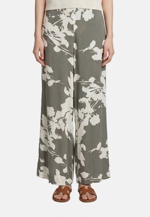 Pantaloni a gamba larga con grande stampa floreale bianca su sfondo oliva, abbinati a sandali color cuoio con punta aperta, mostrati dalla vita ai piedi.