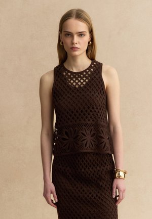 Frau mit glattem blondem Haar trägt ärmelloses braunes Häkeloberteil und passenden Rock, dazu goldene Creolen und Armbänder als Accessoires.