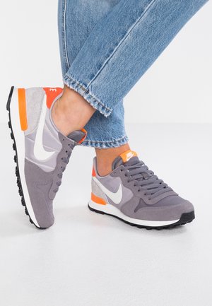 Szare i pomarańczowe buty sportowe Nike noszone z jasnoniebieskimi dżinsami, pokazujące widoki z boku i z góry na białej powierzchni.