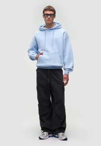 Lyseblå hoodie med kængurulomme, parret med sorte cargo-bukser. Tilbehøret inkluderer chunky hvide sneakers og rektangulære sorte solbriller.