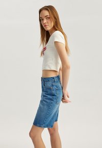 Camiseta corta blanca con logotipo bordado, combinada con una falda de mezclilla azul clara de cintura alta. Textura suave, diseño casual, corte hasta la rodilla.