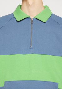 Pull-over zippé en mélange de coton bleu et vert. Présente un col vert et un design à blocs de couleur horizontaux. Texture douce, coupe décontractée.