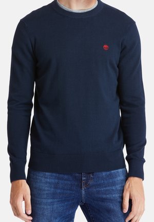 Homme portant un pull col rond bleu marine avec un logo rouge sur la poitrine et un jean bleu, debout les bras détendus le long du corps.