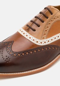 Scarpe eleganti in pelle con pannelli marroni e beige, dettagli brogue, lacci rotondi e una texture liscia con un accento bianco a contrasto.