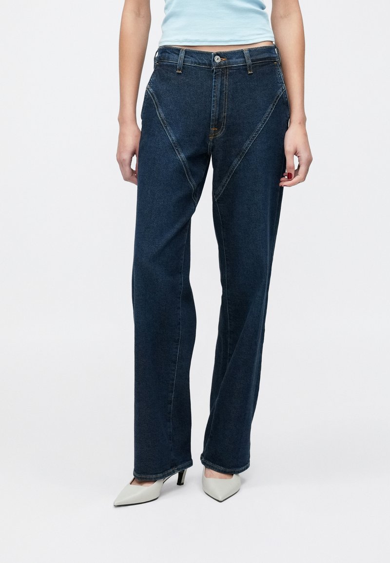 Sötét mosott farmernadrág, laza, egyenes szárú fazonban. Szögletes varrásokkal és kontrasztos cérnavarrással díszítve. Világos színű magassarkúhoz párosítva.