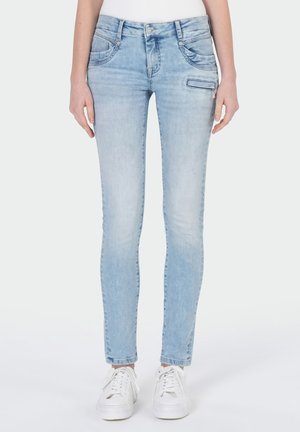 Frau trägt hellblaue Skinny-Jeans mit Reißverschlusstasche an der Seite und weißen Turnschuhen, steht vor einfarbigem Hintergrund.