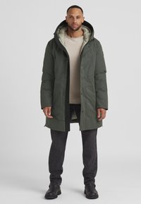 Parka in dunklem Grün, mit Kapuze, Reißverschluss, Druckknöpfen und einem strukturierten Innenfutter. Über einem beigen Pullover und dunklen Jeans getragen.