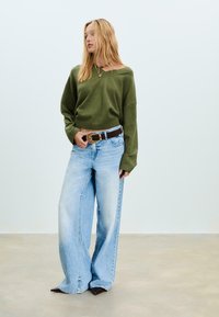 Jonge vrouw met lang blond haar, gekleed in een olijfgroen trui, lichte blauwwijde broek, een bruine riem en puntige donkere schoenen.