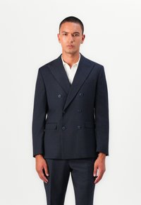 Veste de costume navy double boutonnage en tissu texturé, avec revers en pointe et deux poches avant. Chemise blanche avec col en dessous.