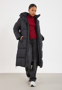 Kvinde iført en lang sort aftensmadjakke, rød rullekravesweater, sorte jeans og hvide sneakers, stående med hænderne i jakke-lommerne.