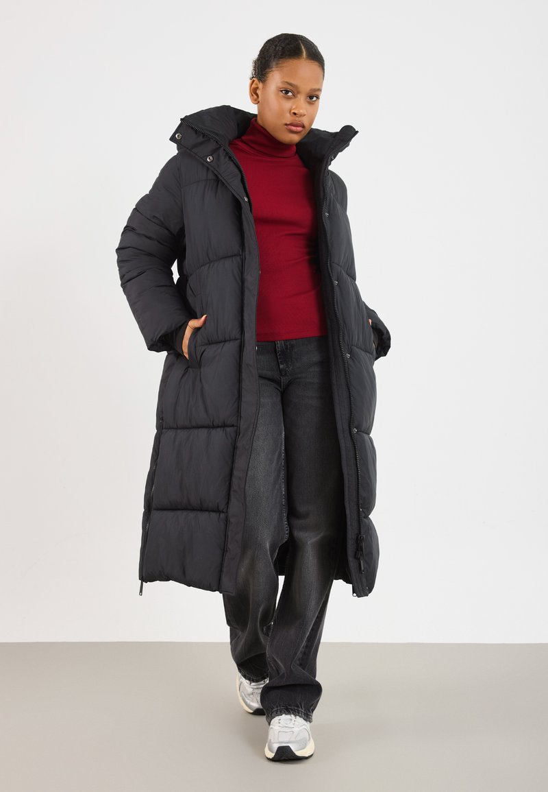 Mulher vestindo um longo casaco puffer preto, suéter de gola alta vermelho, jeans pretos e tênis brancos, em pé com as mãos nos bolsos do casaco.
