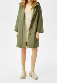 Abrigo largo verde oliva con capucha, dos bolsillos delanteros y cierre de botones; combinado con un vestido de punto crema y zuecos beis con tacón.