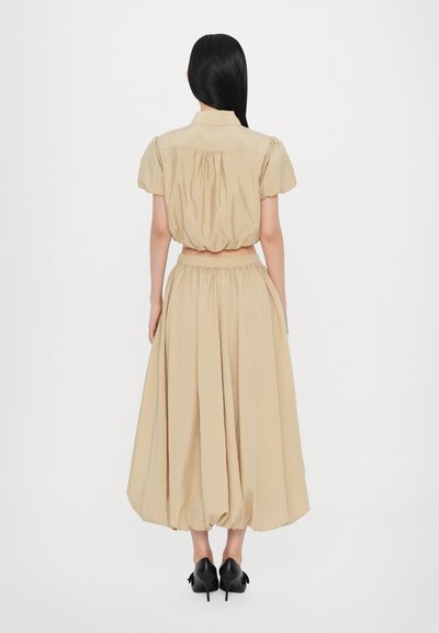 Beiges zweiteiliges Outfit mit einem kurzärmeligen Cropped-Top mit Puffärmeln und einem langen, fließenden, hochgeschnittenen Rock. Getragen mit schwarzen Absätzen.