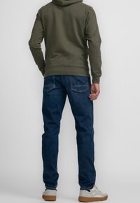 Oliwkowa bluza z kapturem z kieszenią kangurka, zestawiona z niebieskimi dżinsami. Dżinsy mają prosty krój i dwie tylne kieszenie. Białe trampki.