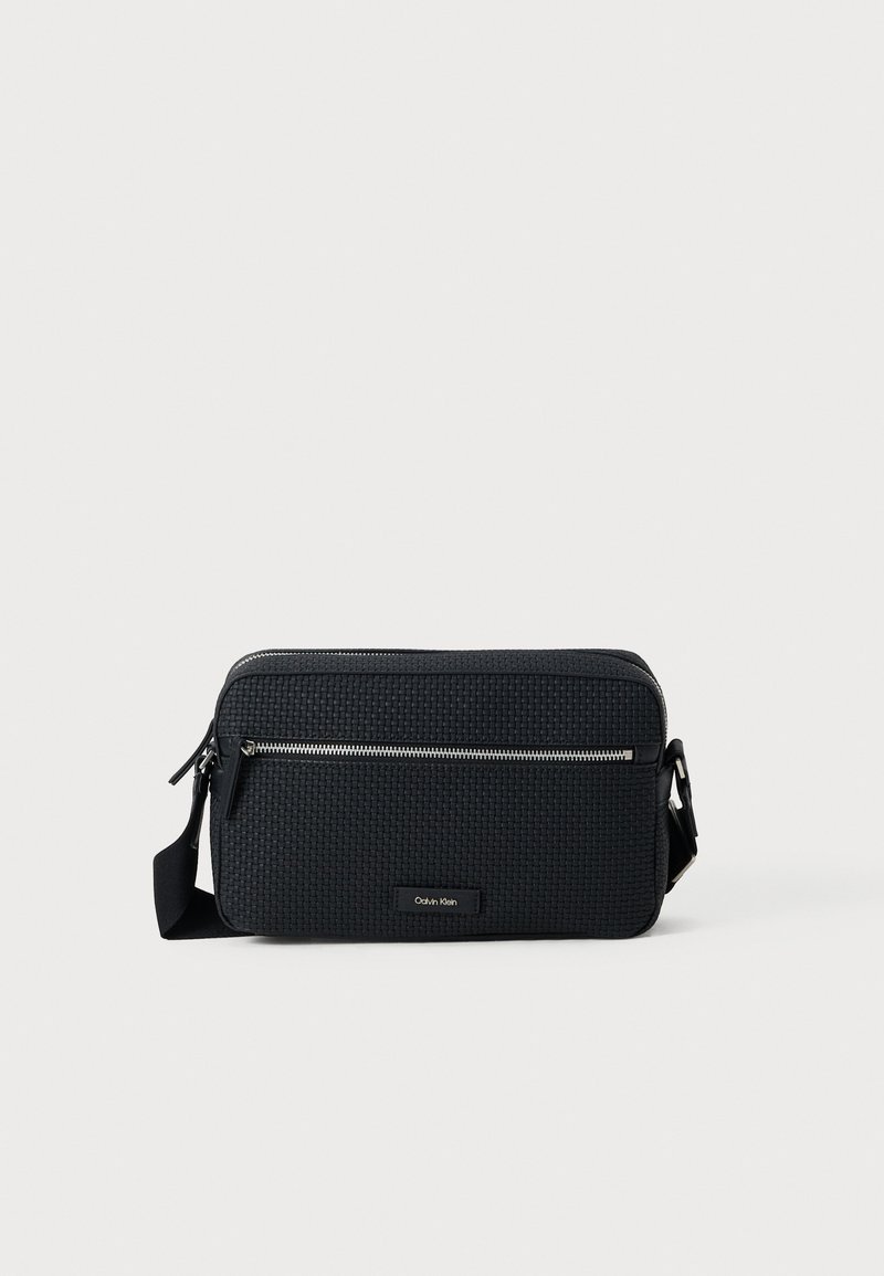 Bolsa transversal tecida em preto, com alça ajustável, fecho zíper e bolso frontal. Apresenta um formato retangular e exterior texturizado. Logotipo da Calvin Klein.