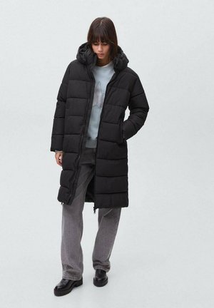 Piumino nero con design trapuntato, cappuccio e taglio lungo. Indossato sopra una felpa azzurra e jeans grigi, abbinato a stivali neri.