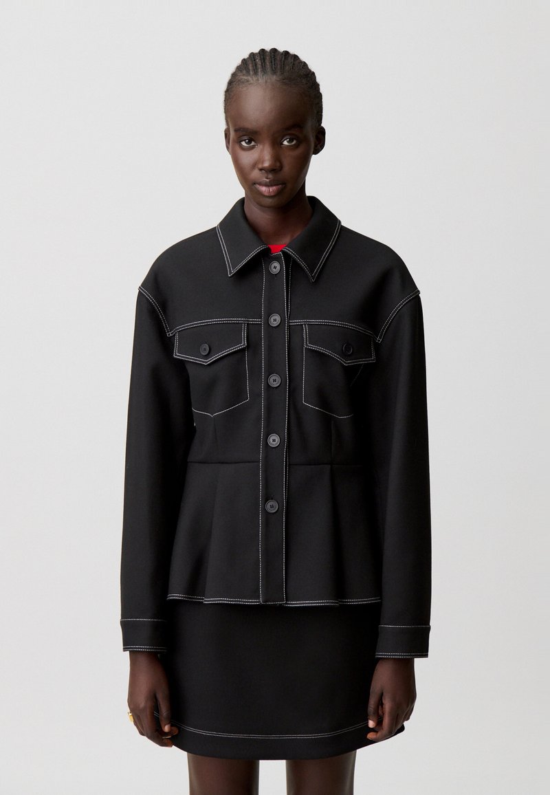 Giacca nera con design a peplo, cuciture bianche a contrasto, due tasche frontali e chiusura con bottoni, indossata sopra una gonna abbinata.